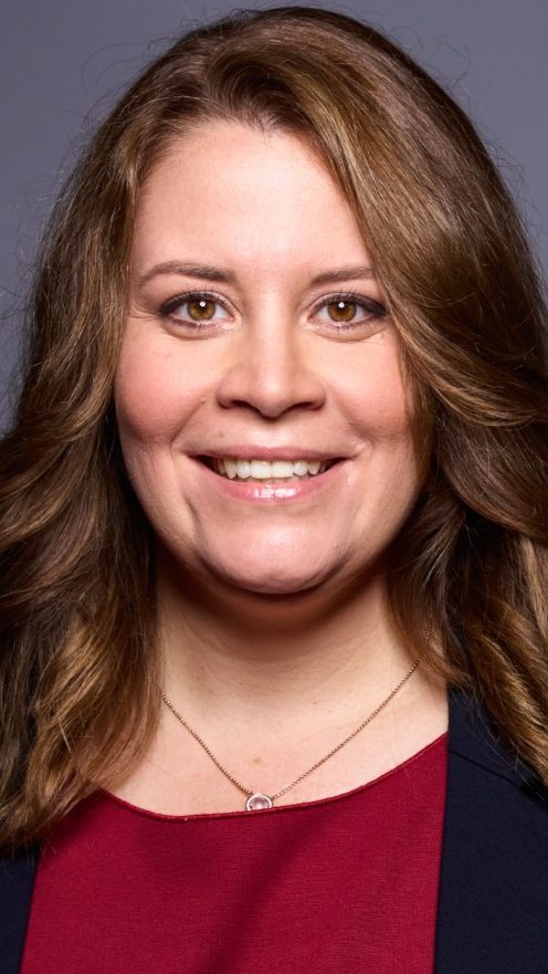 Lächelnde Frau mit langen, lockigen Haaren, trägt eine Jacke und steht im Freien.